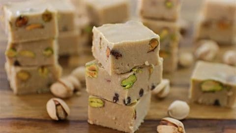 A close-up of several pieces of light brown pistachio fudge stacked on a wooden surface. The fudge is interspersed with whole pistachio nuts, and additional pistachios are scattered around the fudge on the table. The background shows more pieces of fudge.