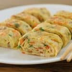 A white plate contains neatly arranged pieces of tamagoyaki, a Japanese rolled omelette, with visible bits of vegetables like carrots and green onions. Chopsticks are placed on the edge of the plate, and the background features a wooden surface.