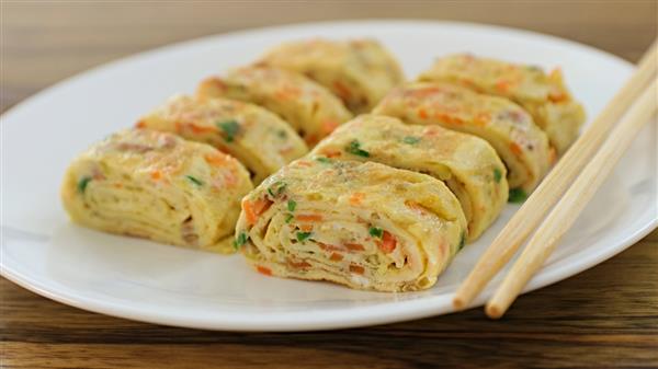 A white plate contains neatly arranged pieces of tamagoyaki, a Japanese rolled omelette, with visible bits of vegetables like carrots and green onions. Chopsticks are placed on the edge of the plate, and the background features a wooden surface.