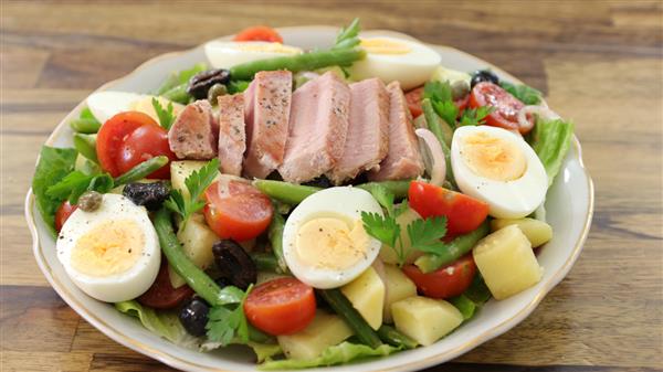 A plate of Nicoise salad featuring sliced seared tuna, hard-boiled eggs, cherry tomatoes, green beans, olives, potatoes, and parsley on a bed of leafy greens. The salad is garnished with capers and arranged on a wooden table.