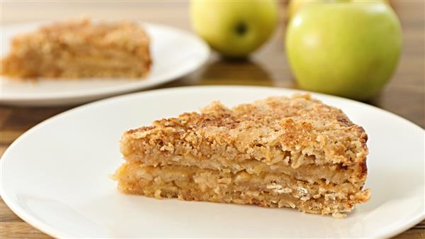 A slice of apple crumb cake sits on a white plate. The cake has a crumbly, golden-brown topping and layered apple filling. In the background, there is a whole cake slice on another plate and two green apples. The scene is set on a wooden surface.