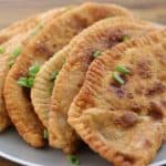 A plate of five crispy, golden-brown, fried dumplings is garnished with chopped green onions and rests on a wooden table. The dumplings are positioned in a semi-circle, highlighting their crimped edges and appetizing appearance.