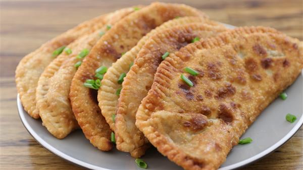 A plate of five crispy, golden-brown, fried dumplings is garnished with chopped green onions and rests on a wooden table. The dumplings are positioned in a semi-circle, highlighting their crimped edges and appetizing appearance.