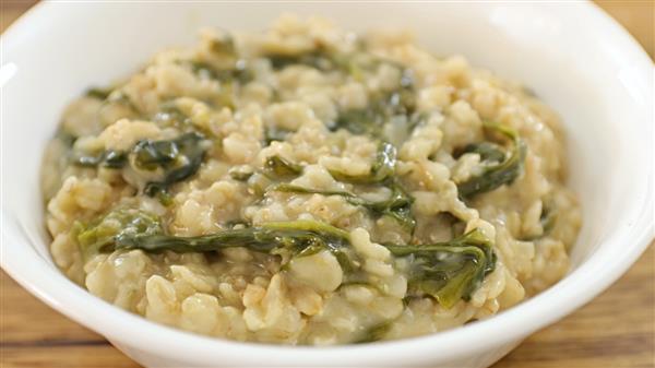 A white bowl filled with a creamy oatmeal dish, mixed with visibly streaked green leafy vegetables. The texture appears hearty and the colors are primarily beige and green, creating a wholesome and nutritious appearance.