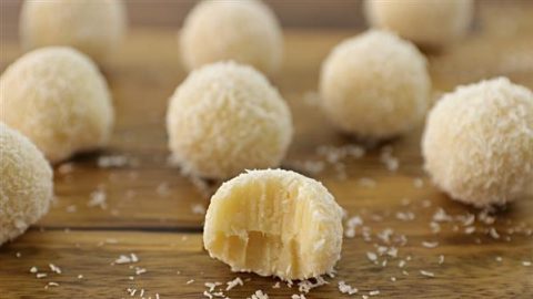 Close-up of white chocolate coconut truffles with one truffle in the foreground having a bite taken out of it. The truffles are coated in shredded coconut and are arranged on a wooden surface, with some coconut shavings scattered around.