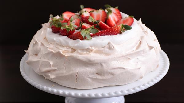 A pavlova topped with whipped cream and halved strawberries sits on a white cake stand. The crisp, light meringue has a slightly cracked surface, giving it a rustic appearance against a dark background.