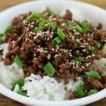 A white bowl filled with white rice topped with cooked ground beef, garnished with sesame seeds and chopped green onions. The bowl is set on a wooden surface.