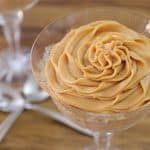 A glass dessert dish containing a creamy pumpkin mousse swirled into a rose shape, sitting on a wooden table. Two silver spoons are placed next to the dish, with another similar dessert dish in the background.