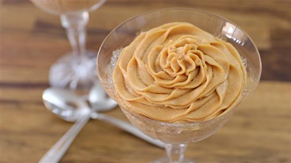 A glass dessert dish containing a creamy pumpkin mousse swirled into a rose shape, sitting on a wooden table. Two silver spoons are placed next to the dish, with another similar dessert dish in the background.