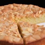 A round almond cake on a white plate with one slice partially removed. The cake has a golden-brown crust and is topped with slivered almonds. It sits against a dark background.