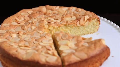 A round almond cake on a white plate with one slice partially removed. The cake has a golden-brown crust and is topped with slivered almonds. It sits against a dark background.