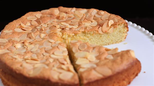 A round almond cake on a white plate with one slice partially removed. The cake has a golden-brown crust and is topped with slivered almonds. It sits against a dark background.