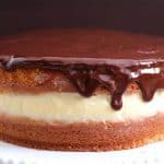 A Boston cream pie with a smooth, shiny chocolate glaze dripping over the sides of the top layer. The cake features a layer of light, creamy filling between two layers of golden, fluffy cake. It is displayed on a white plate with a dark background.