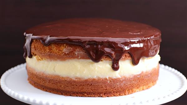 A Boston cream pie with a smooth, shiny chocolate glaze dripping over the sides of the top layer. The cake features a layer of light, creamy filling between two layers of golden, fluffy cake. It is displayed on a white plate with a dark background.