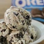 A close-up of a bowl filled with scoops of cookies and cream ice cream, featuring visible chunks of chocolate cookies. In the background, a partly visible Oreo cookie package is out of focus. The ice cream appears creamy and cold, perfect for a treat.