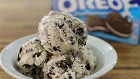 A close-up of a bowl filled with scoops of cookies and cream ice cream, featuring visible chunks of chocolate cookies. In the background, a partly visible Oreo cookie package is out of focus. The ice cream appears creamy and cold, perfect for a treat.