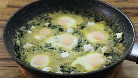 A black frying pan filled with a cooked egg and spinach dish. There are six sunny-side-up eggs nestled in a green spinach mixture, with small white chunks of cheese sprinkled throughout. The pan sits on a wooden surface.