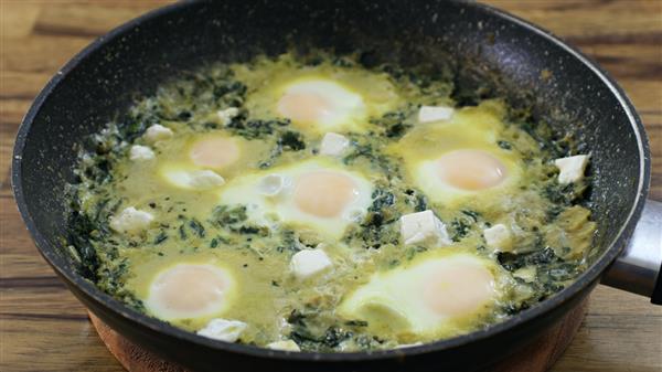 A black frying pan filled with a cooked egg and spinach dish. There are six sunny-side-up eggs nestled in a green spinach mixture, with small white chunks of cheese sprinkled throughout. The pan sits on a wooden surface.