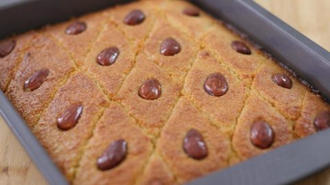 A rectangular tray of freshly baked Basbousa, a Middle Eastern semolina cake, with a golden-brown crust. The cake is cut into diamond-shaped pieces, each topped with a single almond. The cake appears soft and moist with an appetizing texture.