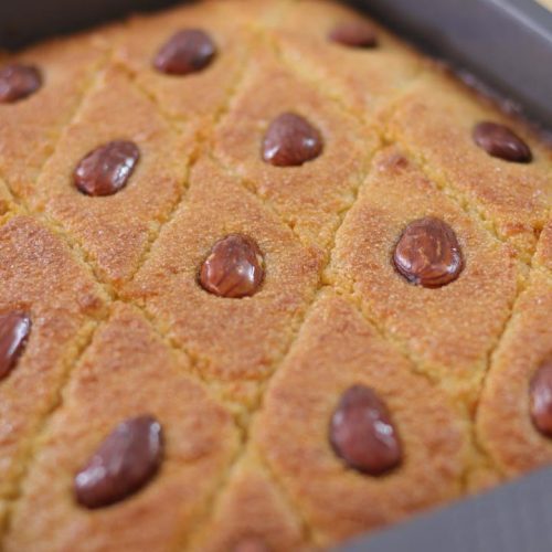 A rectangular tray of freshly baked Basbousa, a Middle Eastern semolina cake, with a golden-brown crust. The cake is cut into diamond-shaped pieces, each topped with a single almond. The cake appears soft and moist with an appetizing texture.