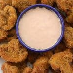 A plate of crispy, breaded fried mushrooms encircles a small blue bowl filled with creamy dipping sauce, placed in the center. The dish is set on a wooden surface, and the mushrooms have a golden-brown color, suggesting they are freshly fried.