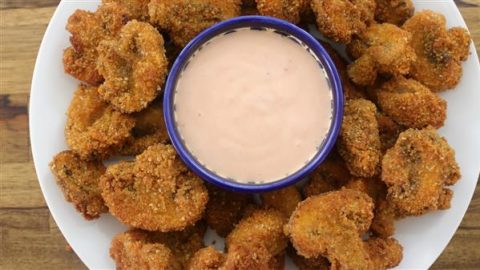 A plate of crispy, breaded fried mushrooms encircles a small blue bowl filled with creamy dipping sauce, placed in the center. The dish is set on a wooden surface, and the mushrooms have a golden-brown color, suggesting they are freshly fried.