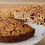 A hand is lifting a slice of a baked oatmeal cake with a serving spatula. The cake is round, appears slightly golden brown, and is dotted with visible raisins or chocolate chips. It is resting on a white plate, and the background shows a wooden surface.
