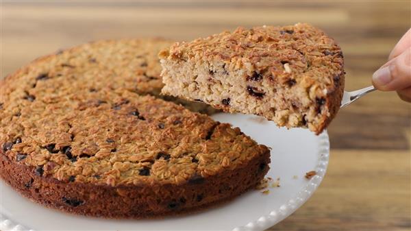 A hand is lifting a slice of a baked oatmeal cake with a serving spatula. The cake is round, appears slightly golden brown, and is dotted with visible raisins or chocolate chips. It is resting on a white plate, and the background shows a wooden surface.