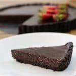 A slice of dark chocolate tart with a chocolate crust sits on a white plate. In the background, the rest of the tart is visible with several strawberries arranged on top. The table underneath is wooden.