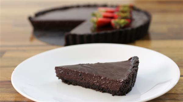 A slice of dark chocolate tart with a chocolate crust sits on a white plate. In the background, the rest of the tart is visible with several strawberries arranged on top. The table underneath is wooden.