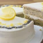 A whole lemon poppy seed cake with white frosting is topped with a lemon slice and garnished with poppy seeds and lemon zest. A slice of the cake is being lifted out with a metal server, showcasing the cake's interior. The background features a wooden surface.