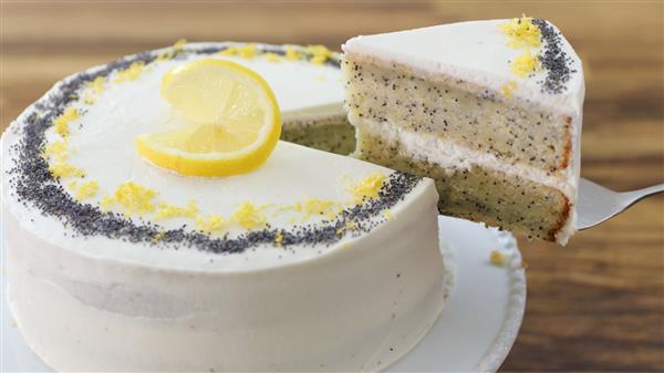 A whole lemon poppy seed cake with white frosting is topped with a lemon slice and garnished with poppy seeds and lemon zest. A slice of the cake is being lifted out with a metal server, showcasing the cake's interior. The background features a wooden surface.