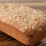 A freshly baked loaf of whole grain bread, topped with oats, cooling on a wire rack. The rustic loaf has a golden-brown crust and is set against a wooden surface background.