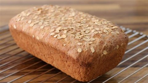 A freshly baked loaf of whole grain bread, topped with oats, cooling on a wire rack. The rustic loaf has a golden-brown crust and is set against a wooden surface background.