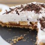 A close-up of a banoffee pie with a slice removed, revealing layers of caramel, sliced bananas, and whipped cream. The pie is topped with chocolate shavings and rests on a white plate. A sliced piece is visible in the background.