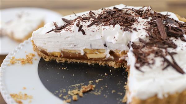 A close-up of a banoffee pie with a slice removed, revealing layers of caramel, sliced bananas, and whipped cream. The pie is topped with chocolate shavings and rests on a white plate. A sliced piece is visible in the background.