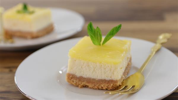 A square slice of layered cheesecake is served on a white plate with a gold fork. The cheesecake has a crumbly crust, a creamy middle layer, and a yellow topping, garnished with a sprig of mint. Another dessert slice is visible in the background.