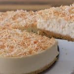 A hand is lifting a slice from a round coconut cheesecake. The cheesecake has a crumbly crust, a creamy filling, and is topped with toasted shredded coconut. It is served on a white plate with a textured edge, with a wooden surface in the background.