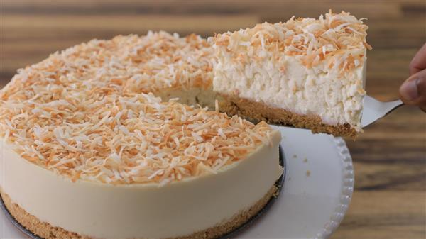 A hand is lifting a slice from a round coconut cheesecake. The cheesecake has a crumbly crust, a creamy filling, and is topped with toasted shredded coconut. It is served on a white plate with a textured edge, with a wooden surface in the background.