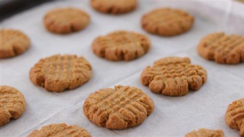 Close-up of rows of freshly baked peanut butter cookies on a baking sheet lined with parchment paper. The cookies have a characteristic crisscross pattern on the tops and are golden brown in color.