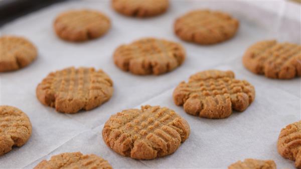 Close-up of rows of freshly baked peanut butter cookies on a baking sheet lined with parchment paper. The cookies have a characteristic crisscross pattern on the tops and are golden brown in color.