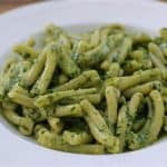 A white plate filled with casarecce pasta coated in green pesto sauce, garnished with finely chopped herbs. The pasta is arranged neatly, and the close-up shot showcases its texture and color. The background is a wooden surface.