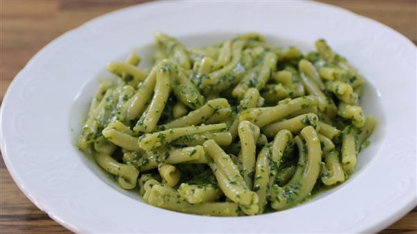 A white plate filled with casarecce pasta coated in green pesto sauce, garnished with finely chopped herbs. The pasta is arranged neatly, and the close-up shot showcases its texture and color. The background is a wooden surface.