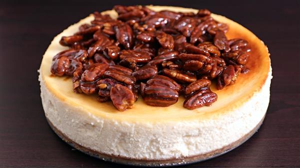 A creamy cheesecake topped with a generous layer of glazed, caramelized pecans sits on a dark surface, highlighting its rich texture and fine, golden crust.