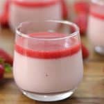 A glass of strawberry mousse topped with a layer of strawberry sauce is shown in the foreground. Additional glasses filled with mousse and several fresh strawberries are visible in the background, all placed on a wooden surface.