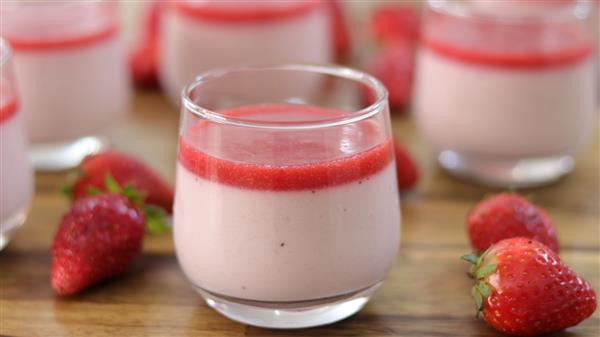 A glass of strawberry mousse topped with a layer of strawberry sauce is shown in the foreground. Additional glasses filled with mousse and several fresh strawberries are visible in the background, all placed on a wooden surface.