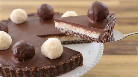 A close-up of a chocolate tart sits on a white plate. The tart features a smooth, glossy chocolate ganache filling, topped with round chocolate and white truffles. A slice is being lifted out with a serving utensil, revealing a layered crust and filling.