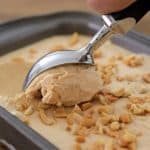 A hand holding an ice cream scooper scoops out a portion of peanut butter ice cream from a rectangular metal container. The ice cream is sprinkled with chopped peanuts. The background shows a wooden surface.