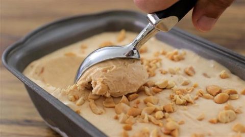 A hand holding an ice cream scooper scoops out a portion of peanut butter ice cream from a rectangular metal container. The ice cream is sprinkled with chopped peanuts. The background shows a wooden surface.