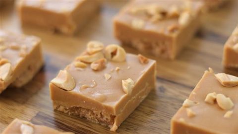 Close-up of square peanut butter fudge pieces arranged on a wooden surface. Each piece has a smooth, creamy texture and is topped with chopped peanuts for added crunch. The fudge looks rich and mouth-watering.